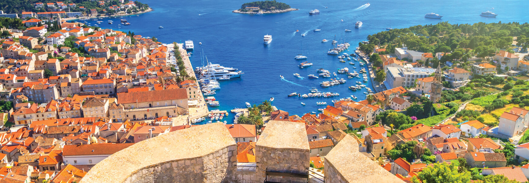 High-angle view from a stone fortification overlooking a Croatian coastal town with red-tiled roofs, a busy harbor, and islands in the sparkling Adriatic Sea.