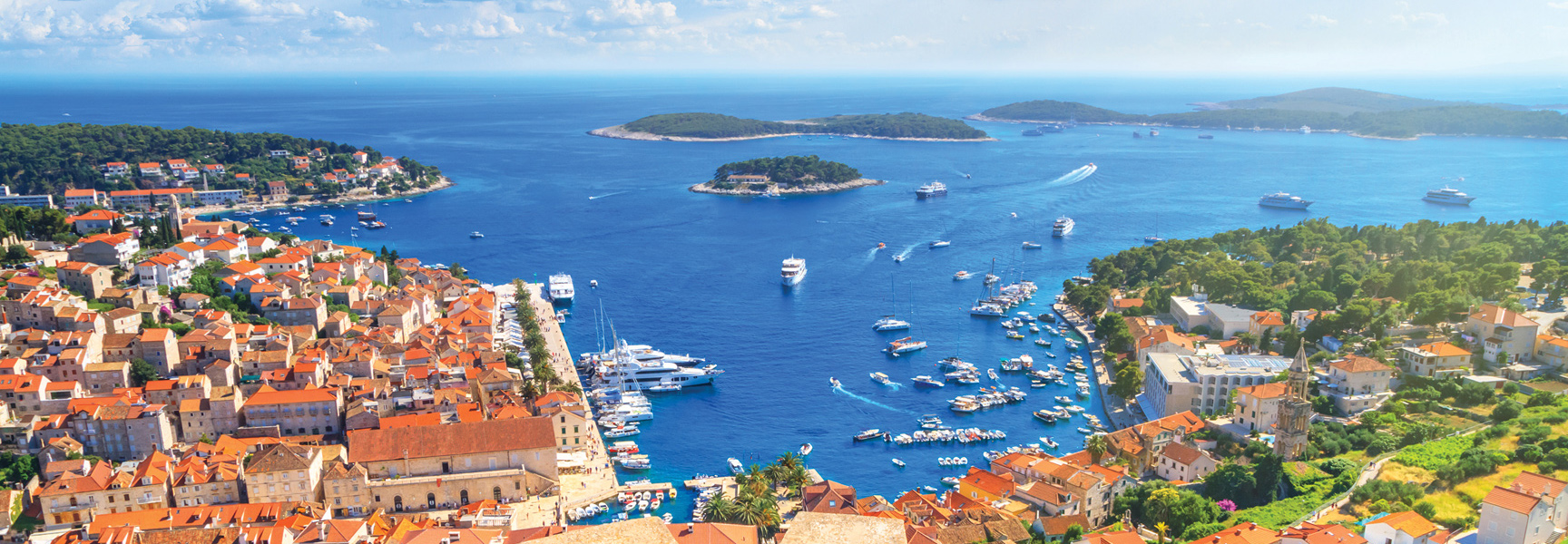 A panoramic, sunny view of a Croatian coastal town with red-roofed buildings, overlooking a bright blue sea filled with boats and several islands.