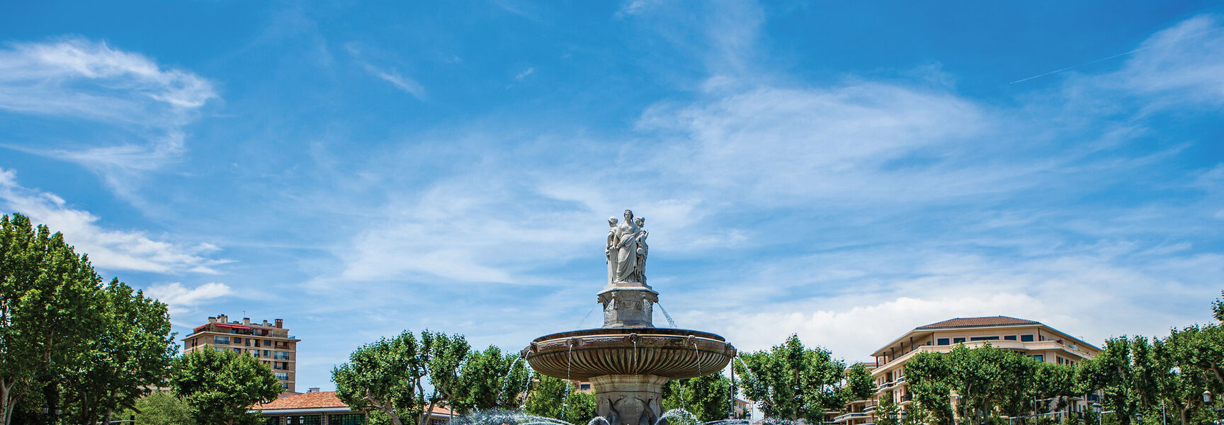 The ornate Fontaine de la Rotonde stands in a sunny city square in Provence, France, surrounded by trees and urban architecture under a blue sky.
