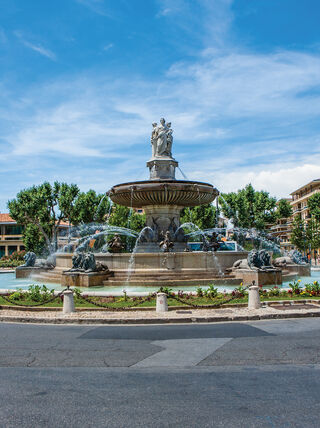 The ornate Fontaine de la Rotonde stands in a sunny city square in Provence, France, surrounded by trees and urban architecture under a blue sky.