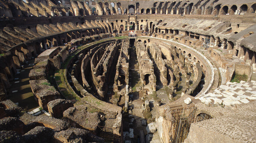 A wide-angle interior view of the ancient Colosseum in Rome, Italy, featuring the exposed stone structures and crumbling tiers of the historic amphitheater.