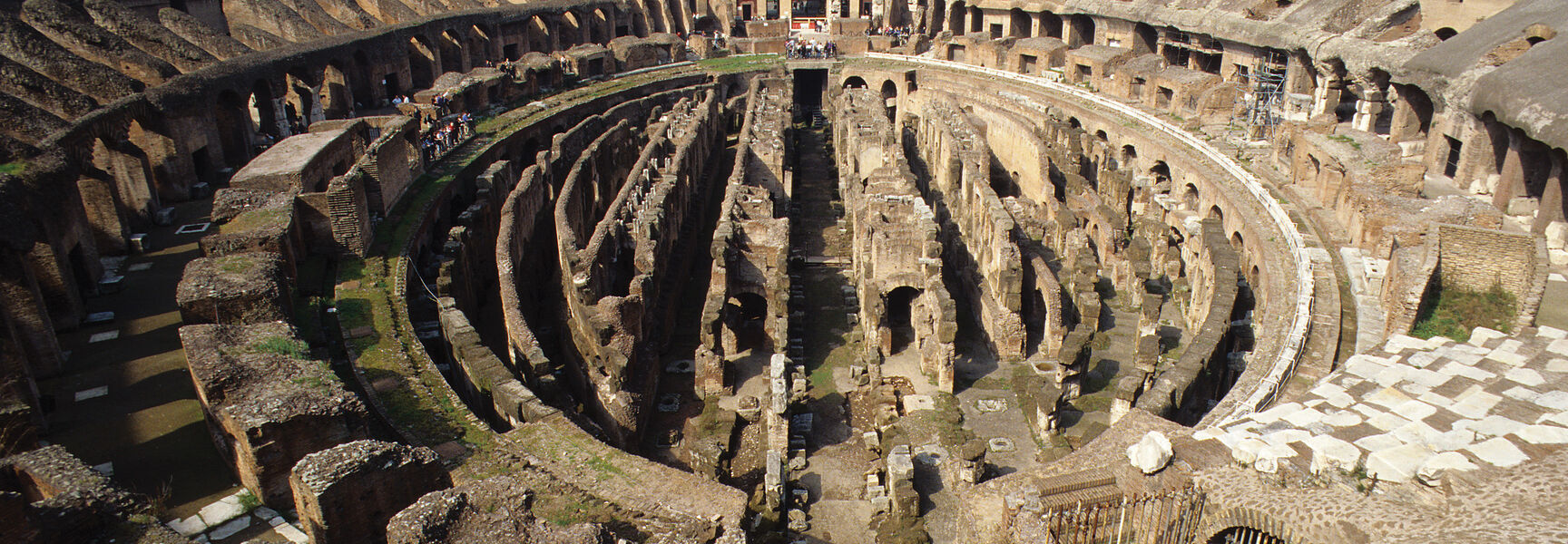 A wide-angle interior view of the ancient Colosseum in Rome, Italy, featuring the exposed stone structures and crumbling tiers of the historic amphitheater.