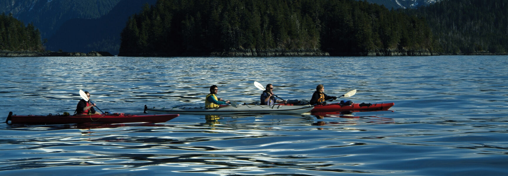 People paddling kayaks on calm water in Southeast Alaska with snow-capped mountains and evergreen forests in the background.