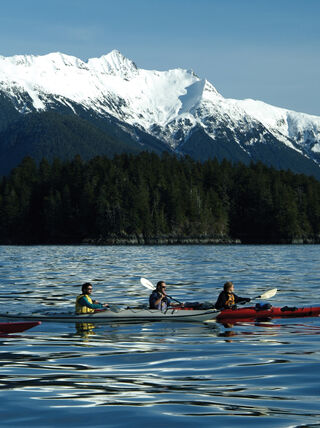 People paddling kayaks on calm water in Southeast Alaska with snow-capped mountains and evergreen forests in the background.