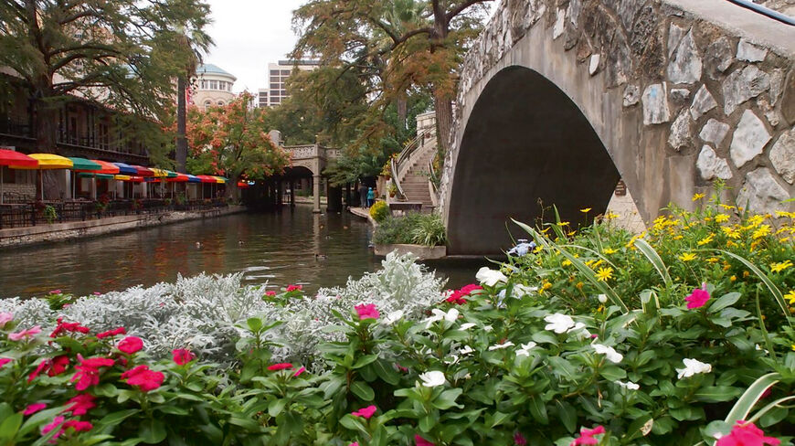 Colorful flowers in the foreground lead to a stone arched bridge over the San Antonio River Walk in Texas, lined with vibrant cafe umbrellas.