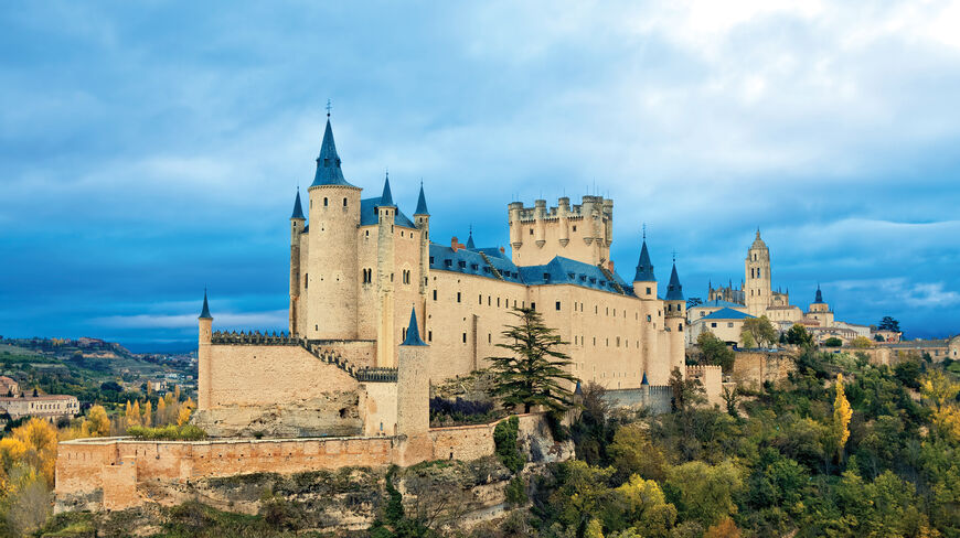 The Alcázar of Segovia, a historic medieval castle in Spain, stands on a rocky hilltop surrounded by autumn trees under a cloudy blue sky.