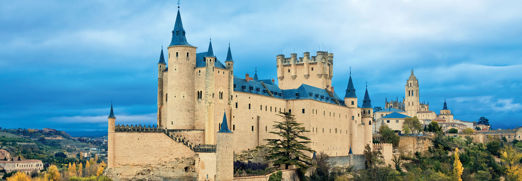 The Alcázar of Segovia, a historic medieval castle in Spain, stands on a rocky hilltop surrounded by autumn trees under a cloudy blue sky.