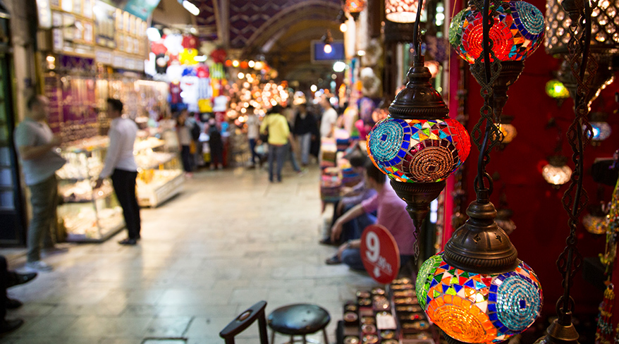 Colorful Turkish mosaic lanterns hang in the foreground of a busy aisle in the Grand Bazaar in Istanbul, Turkey.