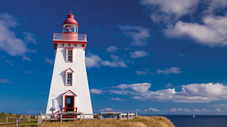 A historic white and red lighthouse stands on a grassy cliff overlooking the ocean in Prince Edward Island.