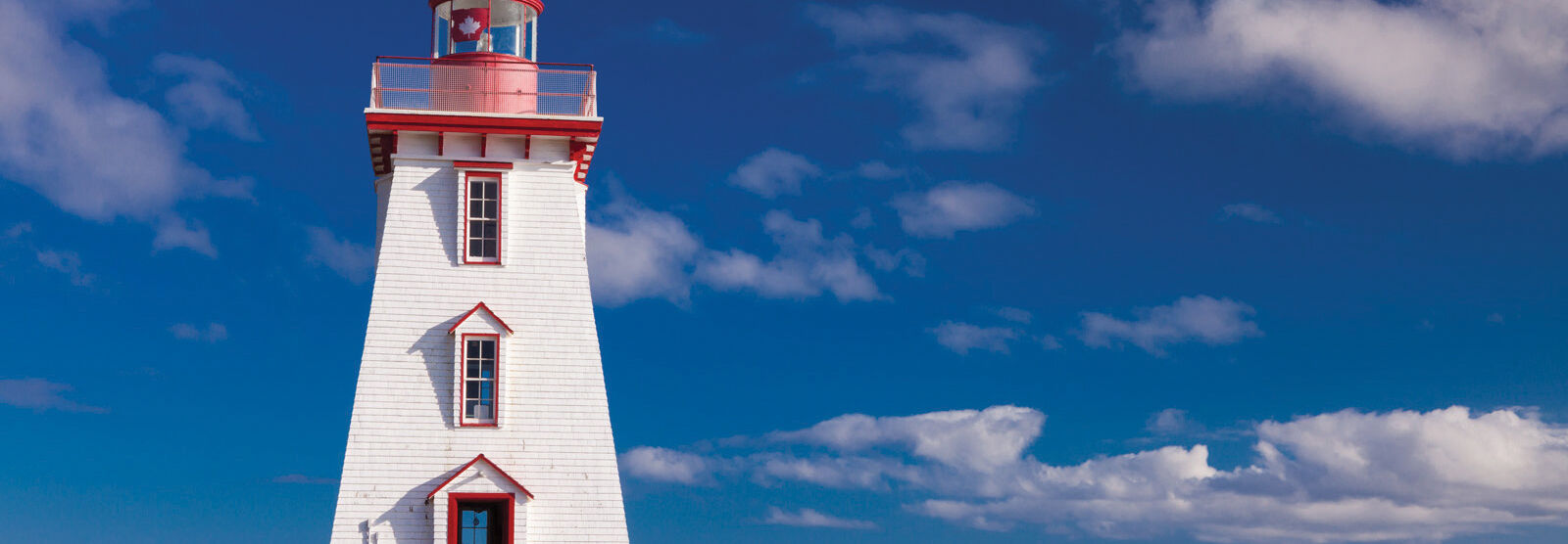 A historic white and red lighthouse stands on a grassy cliff overlooking the ocean in Prince Edward Island.