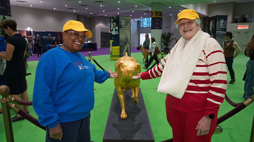 Two women in yellow hats pose with a golden dog statue at the Westminster Kennel Club Dog Show in the Javitz Center, New York.