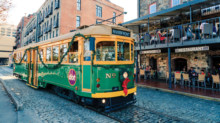 A festive green and yellow dot trolley decorated with Christmas garland travels along a cobblestone street in the historic riverfront district of Savannah, Georgia.