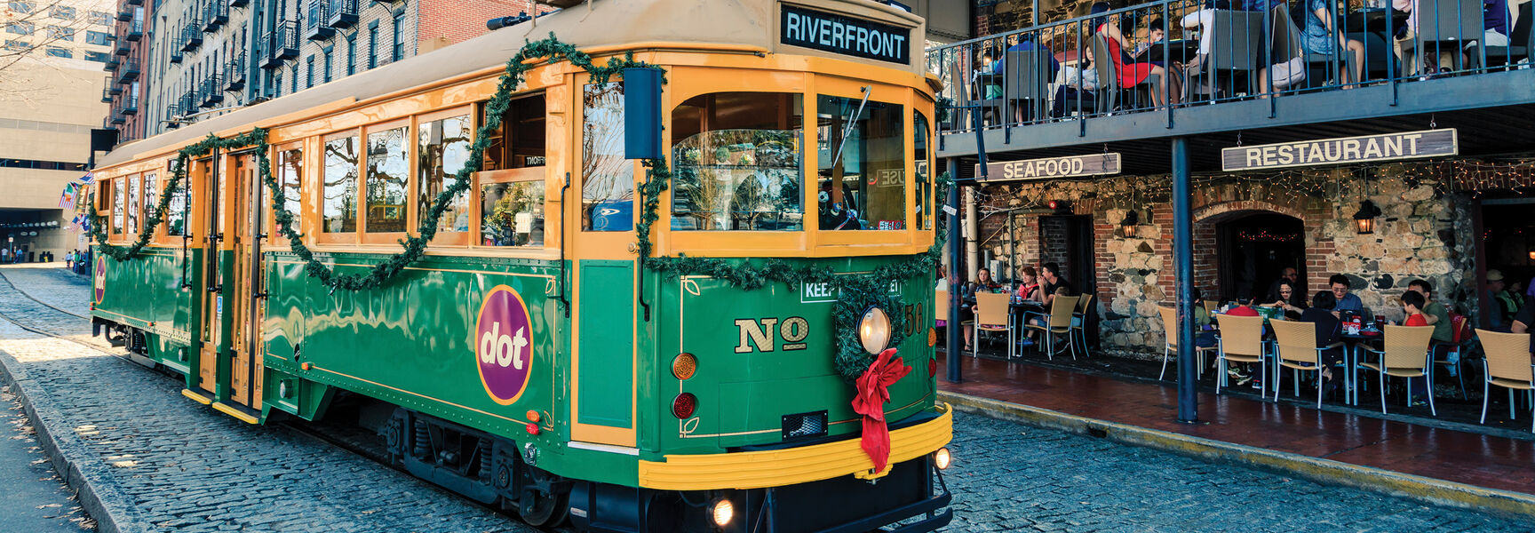 A festive green and yellow dot trolley decorated with Christmas garland travels along a cobblestone street in the historic riverfront district of Savannah, Georgia.