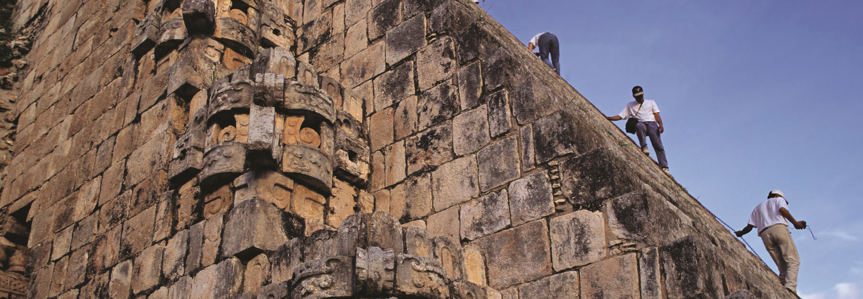 Archaeologists climb the steep, stone side of a large, ancient Maya pyramid with carved details in Mexico.