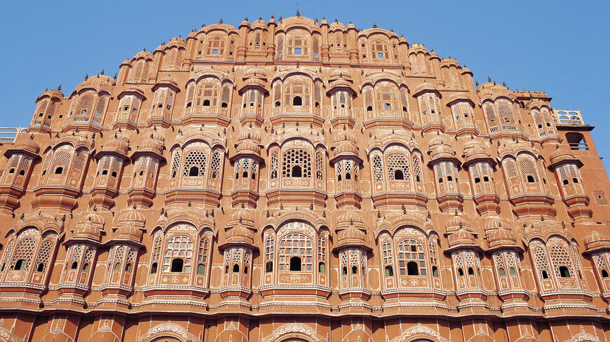 The intricate red and pink sandstone facade of the Hawa Mahal in Jaipur, India, featuring numerous ornate windows under a clear blue sky.