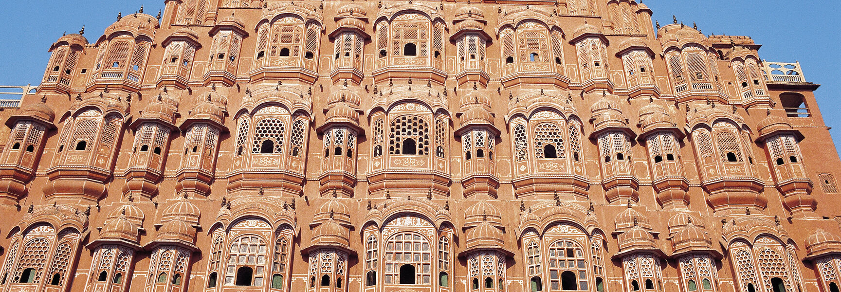 The intricate red and pink sandstone facade of the Hawa Mahal in Jaipur, India, featuring numerous ornate windows under a clear blue sky.