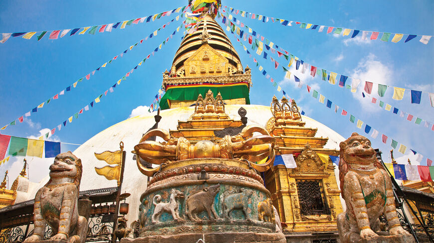 A grand Buddhist temple in Nepal featuring a large white stupa adorned with colorful prayer flags and flanked by ancient stone lion statues.