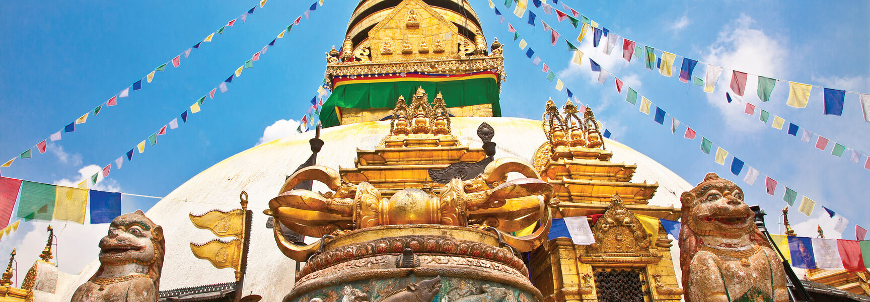 A grand Buddhist temple in Nepal featuring a large white stupa adorned with colorful prayer flags and flanked by ancient stone lion statues.