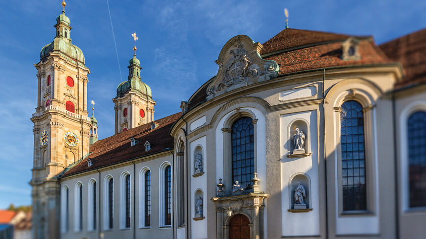 The ornate Baroque facade and twin green-domed towers of the Abbey of Saint Gall stand against a clear blue sky in Switzerland.