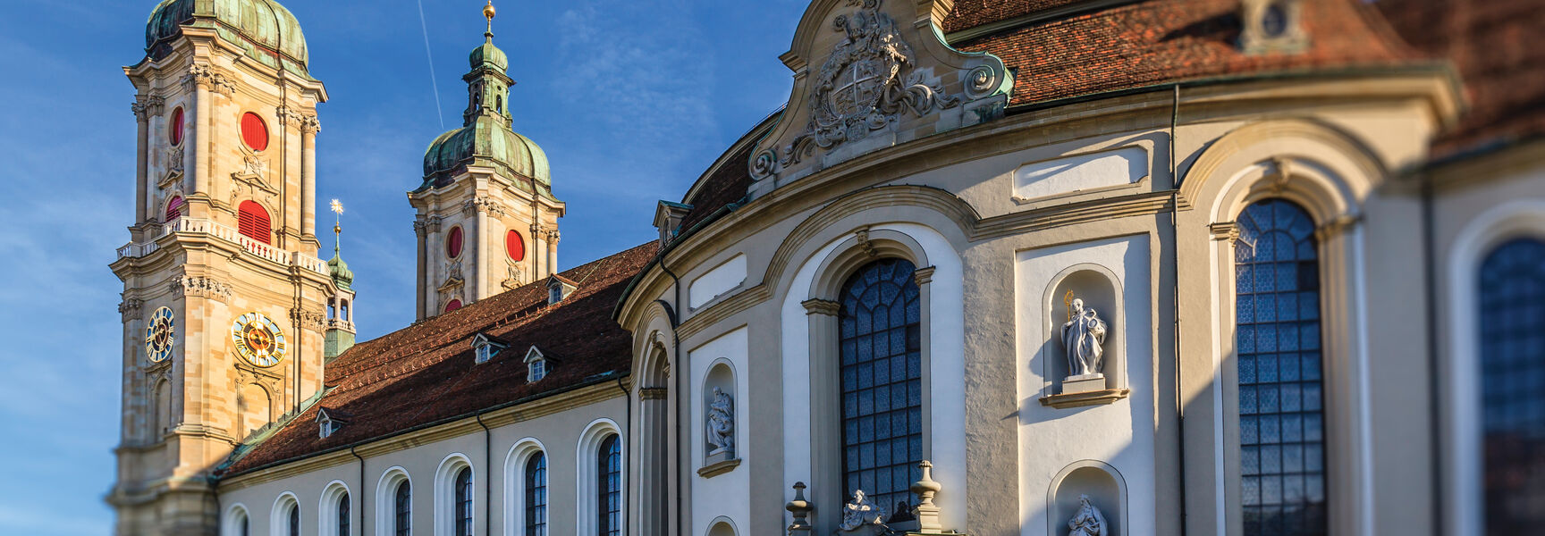 The ornate Baroque facade and twin green-domed towers of the Abbey of Saint Gall stand against a clear blue sky in Switzerland.