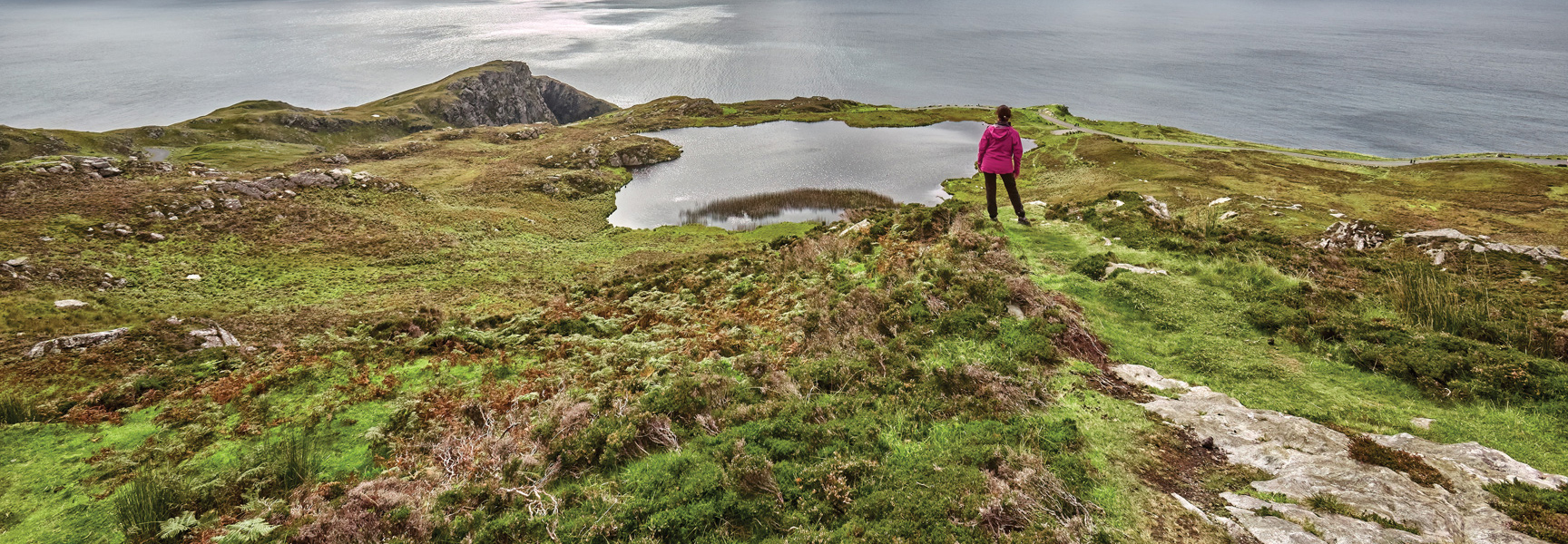 A hiker in a pink jacket stands on a green, mossy cliff overlooking a lake and the ocean in coastal Ireland.