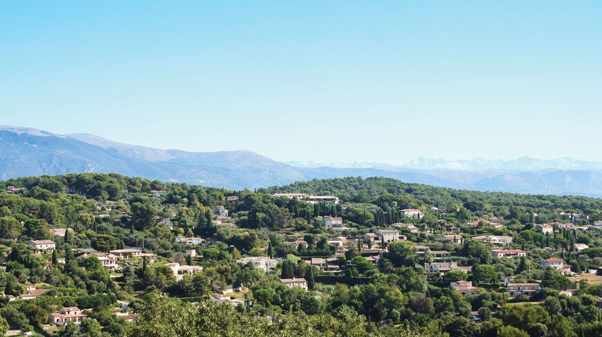 A panoramic view of houses nestled in lush green trees on the French Riviera near Nice, with mountains under a clear blue sky.