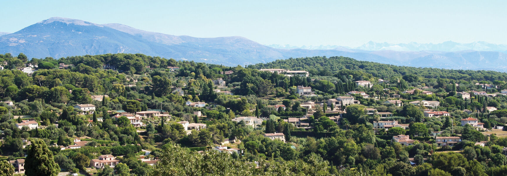 A panoramic view of houses nestled in lush green trees on the French Riviera near Nice, with mountains under a clear blue sky.