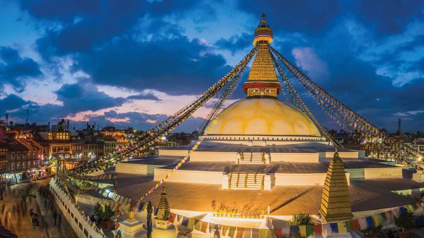 An illuminated Buddhist stupa in the Kathmandu Valley, Nepal, stands under a twilight sky, adorned with colorful prayer flags and surrounded by city buildings.