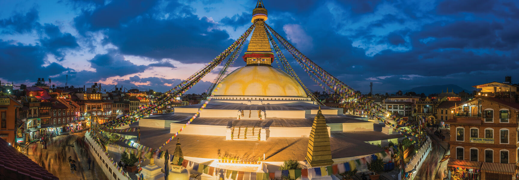 An illuminated Buddhist stupa in the Kathmandu Valley, Nepal, stands under a twilight sky, adorned with colorful prayer flags and surrounded by city buildings.