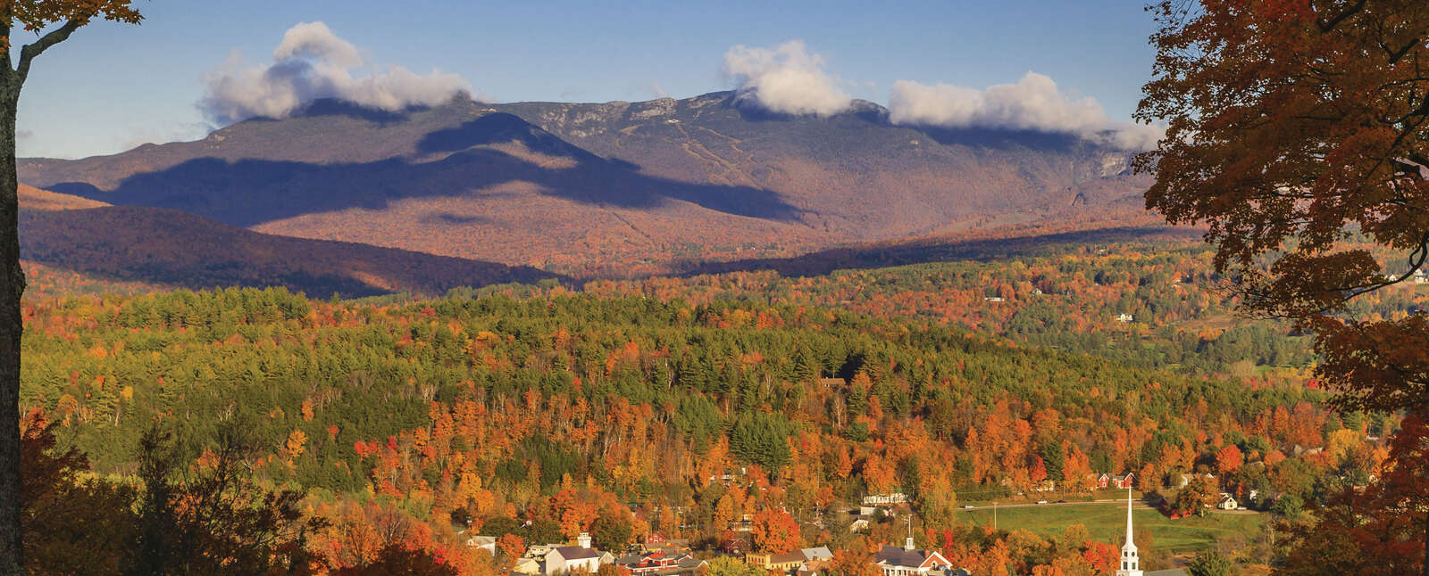 A picturesque autumn view of Vermont's Green Mountain State woodlands surrounding a small town with a white steeple nestled below mist-covered mountains.