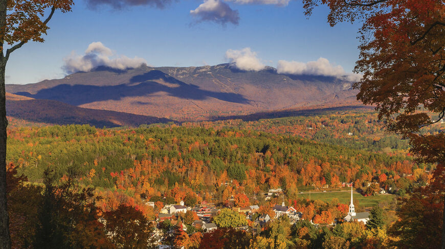 A picturesque autumn view of Vermont's Green Mountain State woodlands surrounding a small town with a white steeple nestled below mist-covered mountains.