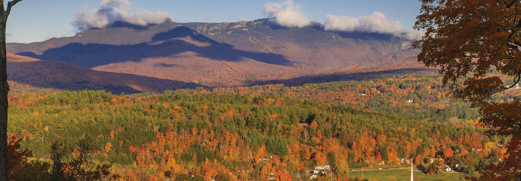 A picturesque autumn view of Vermont's Green Mountain State woodlands surrounding a small town with a white steeple nestled below mist-covered mountains.