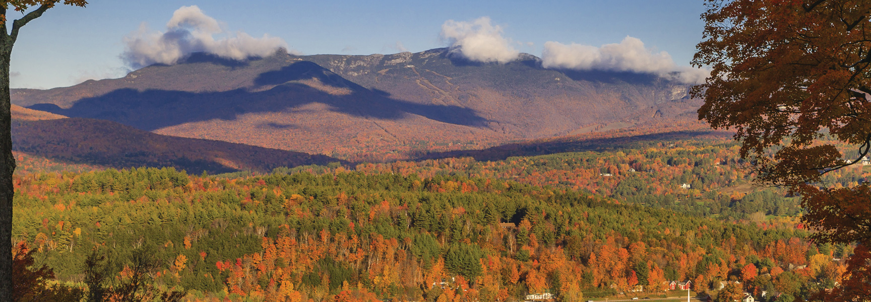 A scenic view of Vermont's mountains and rolling hills covered in vibrant red, orange, and green autumn foliage under a blue sky.