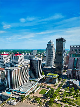 An aerial daytime view of the Louisville skyline, featuring modern skyscrapers and a small urban park under a bright blue sky.