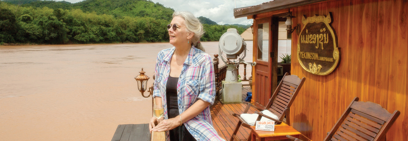 A smiling woman with a glass of wine relaxes on the deck of a wooden riverboat in Vietnam, Laos, or Cambodia.