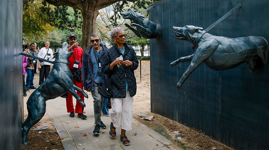 A group of people on a Civil Rights tour in Birmingham, Alabama, view an outdoor sculpture of dogs lunging from a wall.