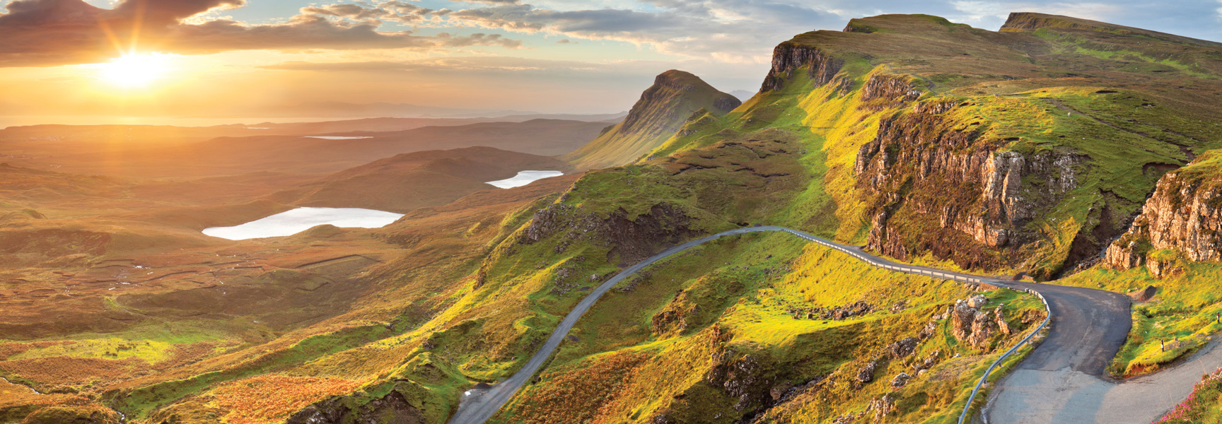 A winding road travels through the dramatic, green mountains of the Scottish Highlands as the sun shines over distant lochs.