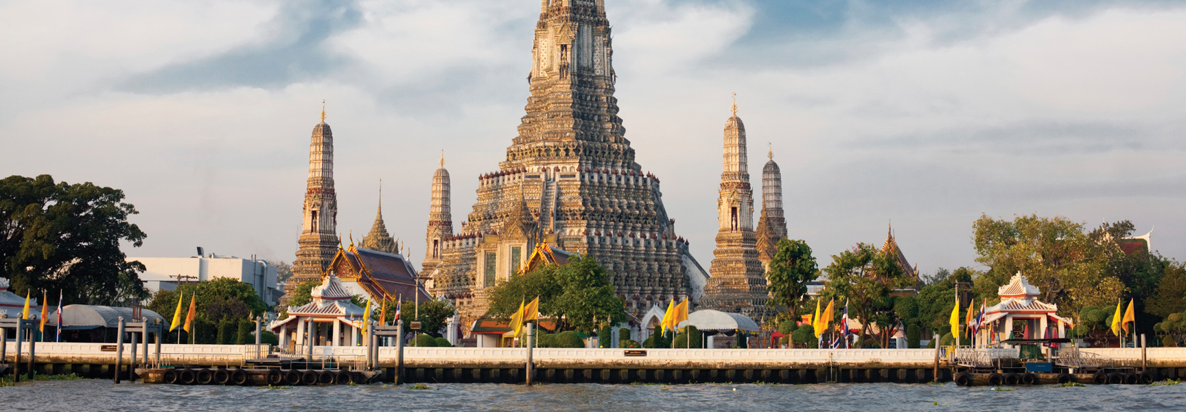 A grand Buddhist temple with towering spires stands on the riverbank in Thailand under a partly cloudy sky.