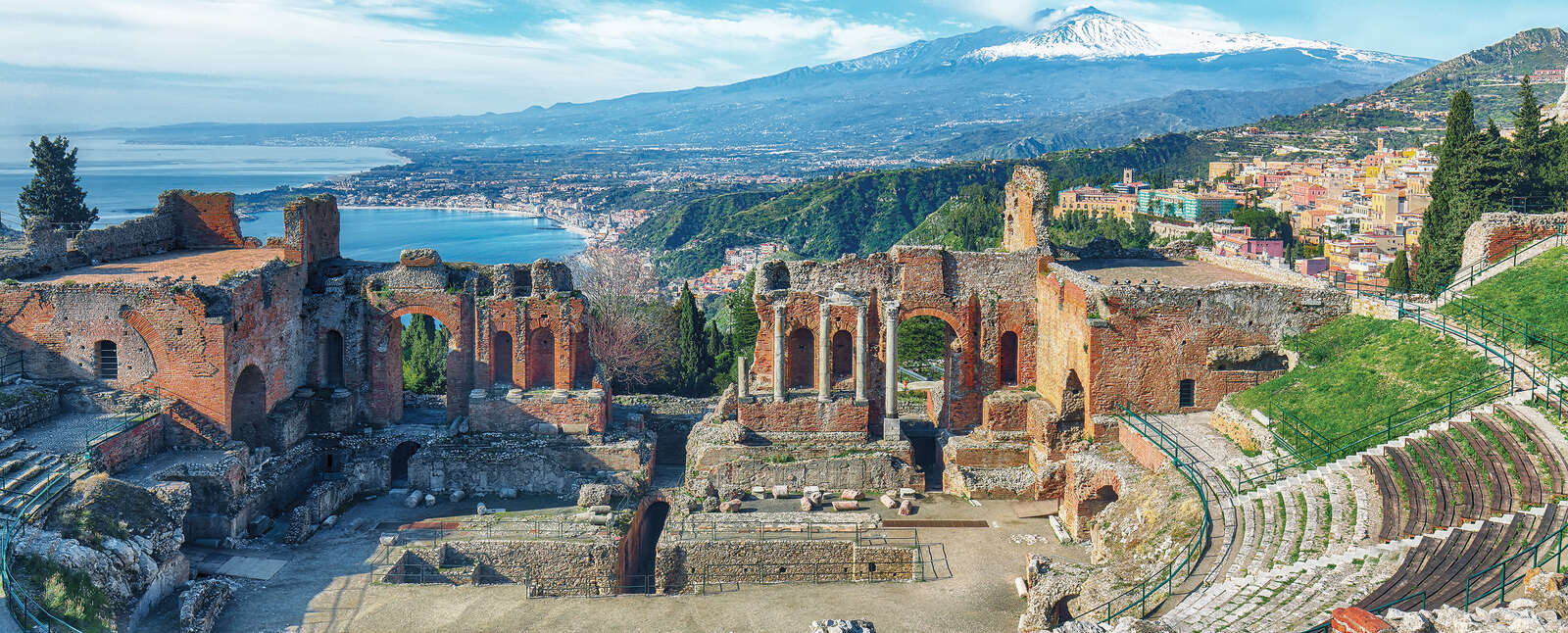 Ancient Roman and Greek ruins of the theatre in Taormina, Sicily, overlooking the coastline with a snow-capped Mount Etna in the background.