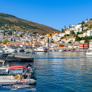 Sunlight shines on the harbor of Hydra, Greece, where boats are docked near a hillside village with white buildings and orange-tiled roofs.