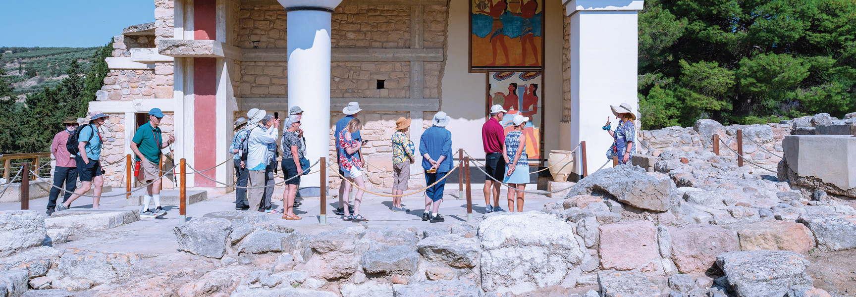 A group of tourists listens to a guide while standing among the ancient ruins and colorful frescoes at the Palace of Knossos in Crete, Greece.
