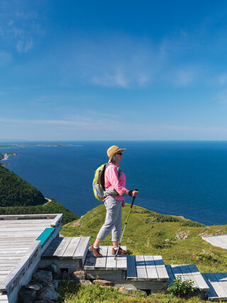 A hiker with a backpack stands on a wooden boardwalk overlooking the scenic coastal mountains and blue ocean of the Nova Scotia Highlands.