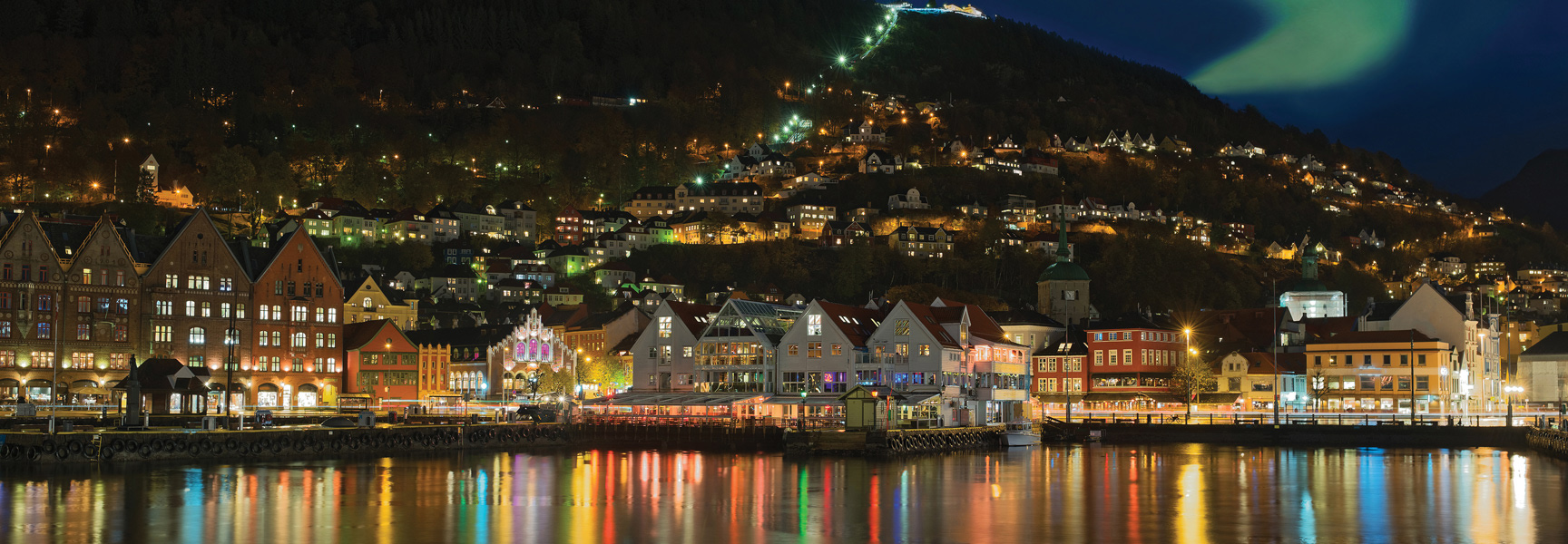The colorful, historic buildings of Bergen, Norway are lit up at night along the waterfront, with the aurora borealis faintly visible overhead.