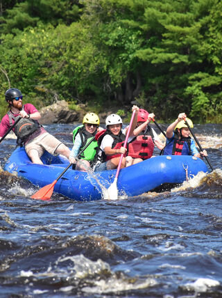 A group whitewater rafting in a blue raft through the rapids of Sandstone, Minnesota, surrounded by the dense green trees of the Northwoods.
