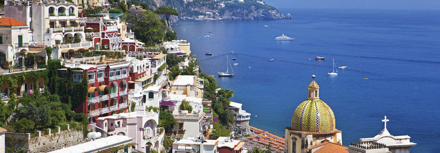 A panoramic view of the colorful cliffside villages of the Amalfi Coast in Italy overlooking the deep blue sea dotted with boats.