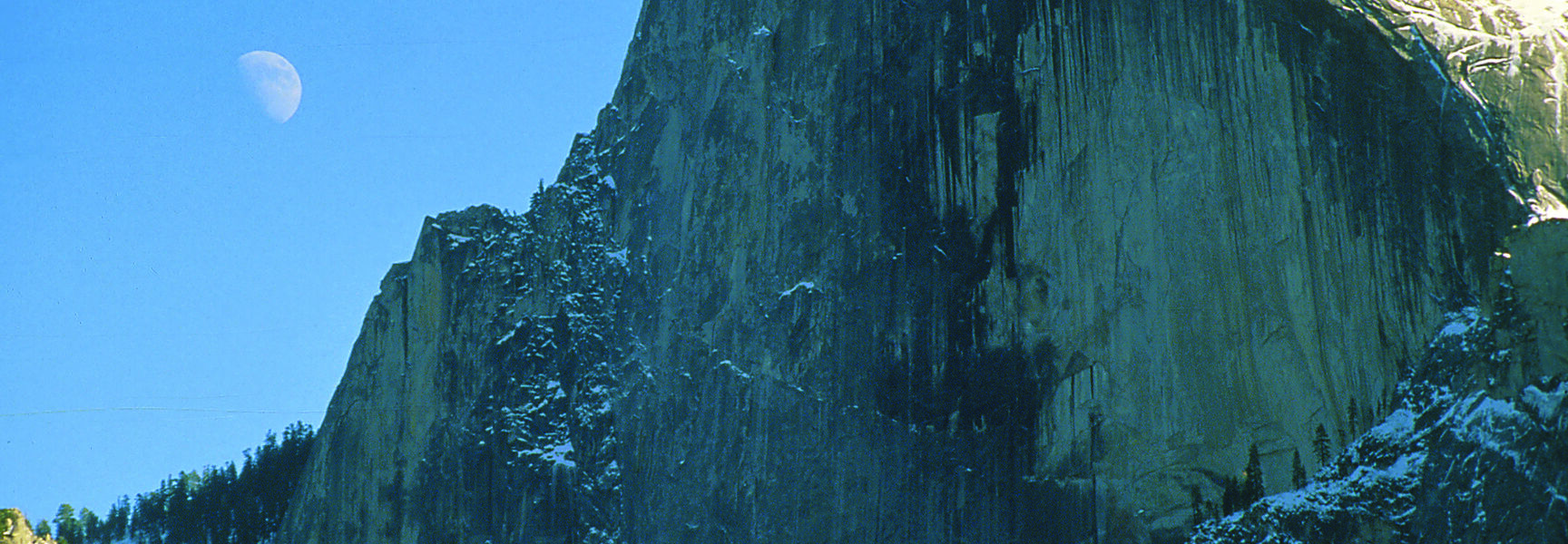 The granite face of Half Dome in Yosemite, California, stands under a clear blue sky with a crescent moon in the distance.