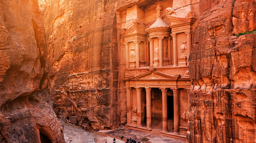 The ancient Treasury at Petra, known as Al-Khazneh, is seen carved into a red rock canyon and lit by warm sunlight.