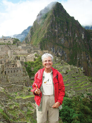 A smiling woman in a red jacket stands before the ancient stone ruins and terraced hillsides of Machu Picchu in Peru.