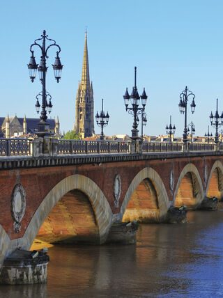 The historic Pont de Pierre bridge spans across a river in Bordeaux, France, featuring ornate lampposts and a cathedral spire in the distance.