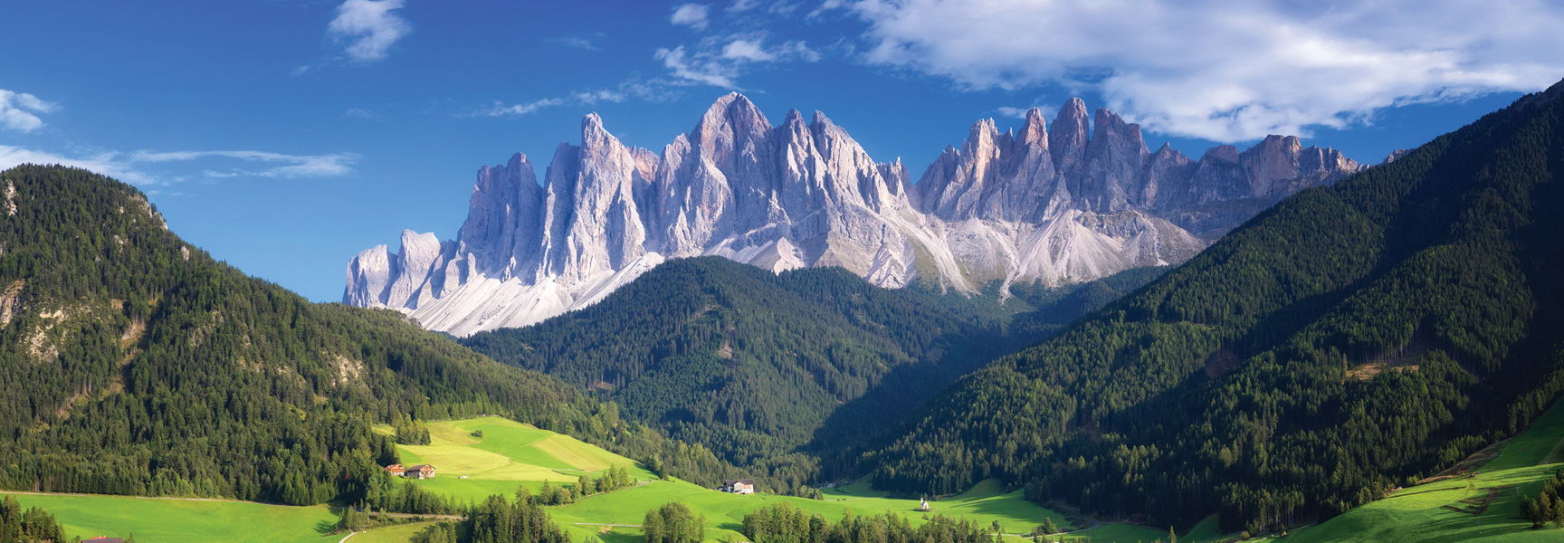 The jagged peaks of the Dolomites mountain range in Italy rise above lush green valleys and dense forests under a partly cloudy blue sky.